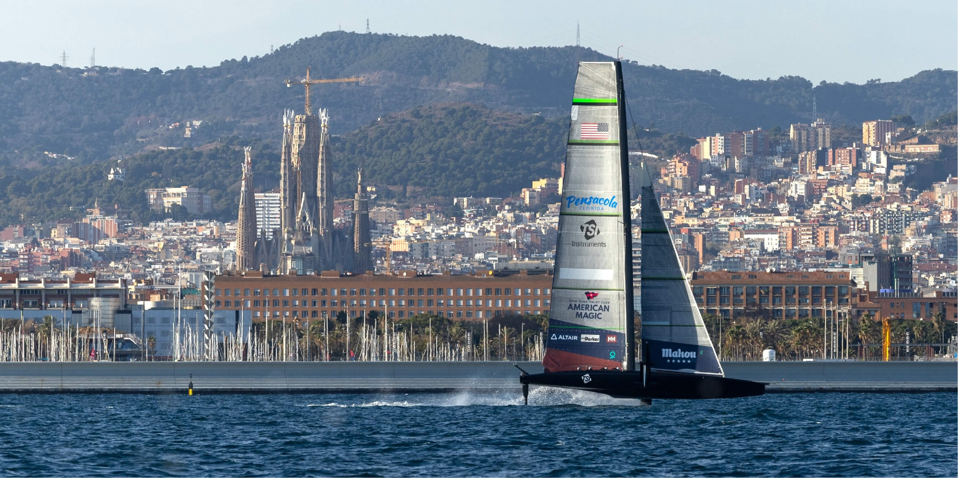 American Magic boat sailing off the coast of Barcelona, with the Sagrada Família in the background.